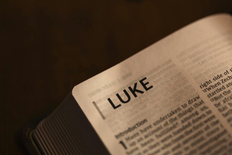Close-up of an open Bible showing the start of the Book of Luke, softly lit on a dark background.