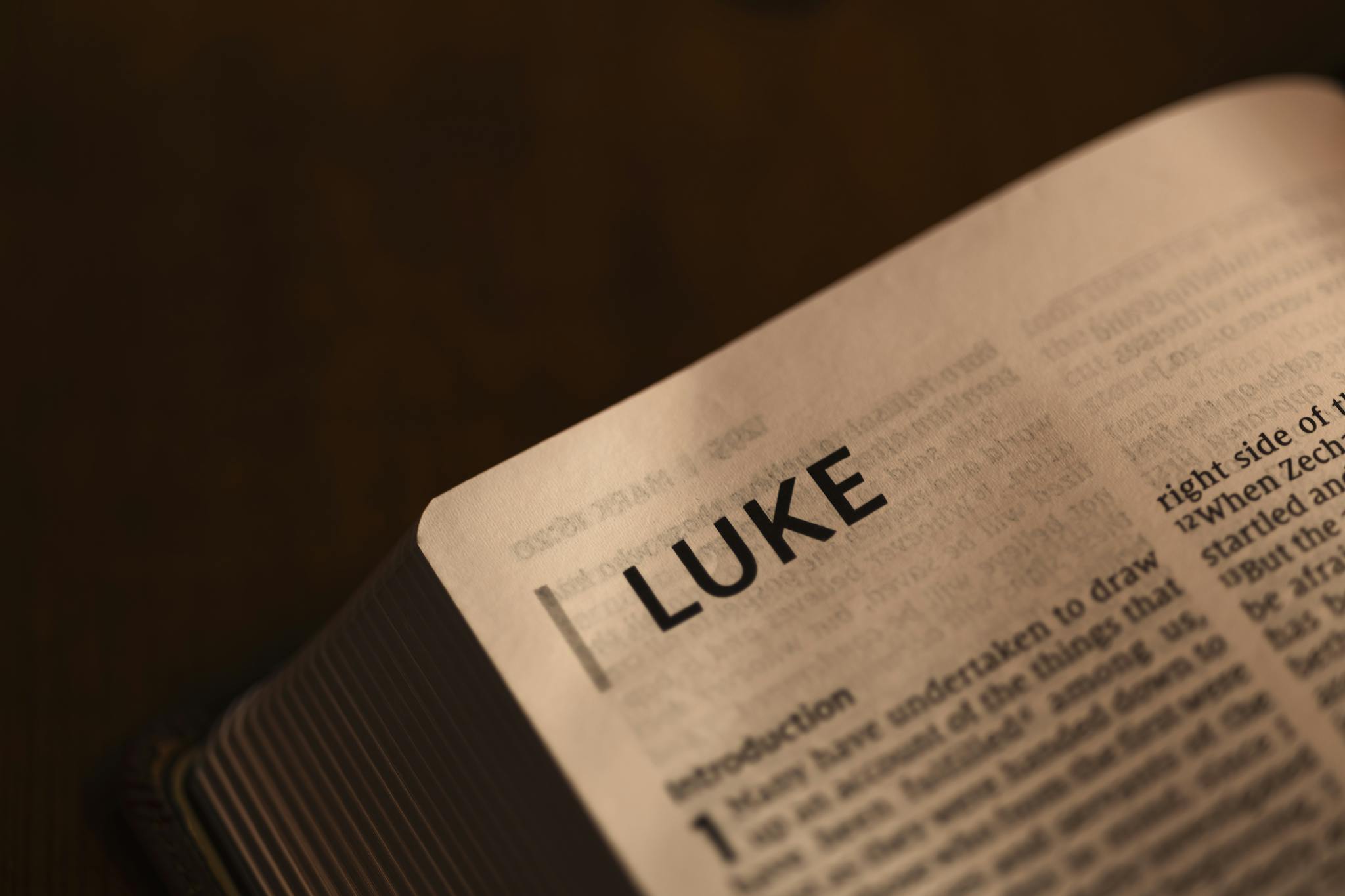 Close-up of an open Bible showing the start of the Book of Luke, softly lit on a dark background.