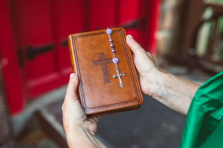 Hands holding a leather-bound Bible with rosary beads, in front of a red door, symbolizing faith.